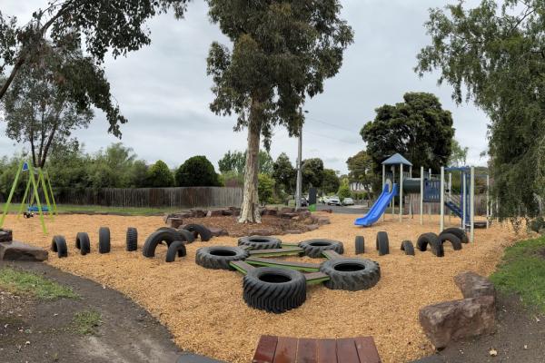 A suburban playground with native trees dotted around, an obstacle course made from tyres, a swing set and a blue slide. Cars and houses can be seen in the distance.